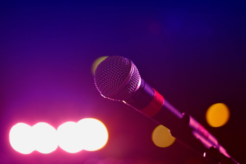 Perspectief van een persoon die karaoke zingt aan een microfoon, met sfeerverlichting en publiek op de achtergrond.