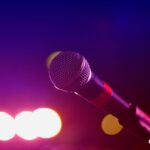 Perspectief van een persoon die karaoke zingt aan een microfoon, met sfeerverlichting en publiek op de achtergrond.
